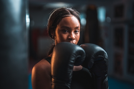 Selective focus photo of an individual in a darkened room wearing black boxing gloves, holding their hands up in a self-protection position.